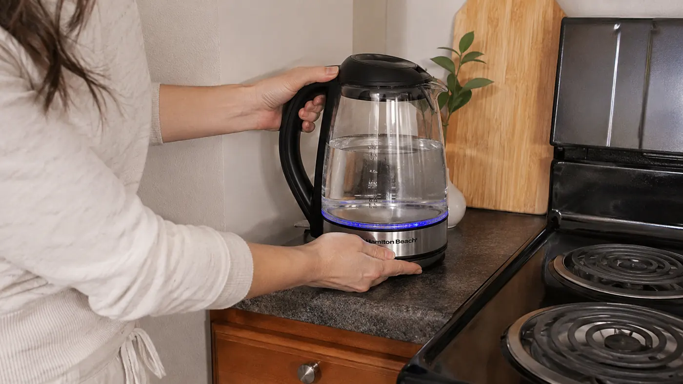 Hand setting the Hamilton Beach electric kettle onto a cleared strip of counter next to a two-burner stove