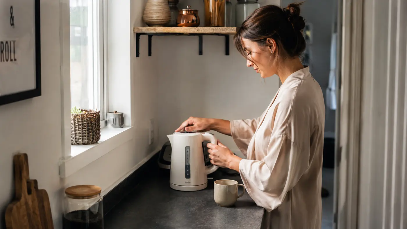 White Hamilton Beach electric kettle sitting on a narrow apartment kitchen counter next to a mug and a tea tin